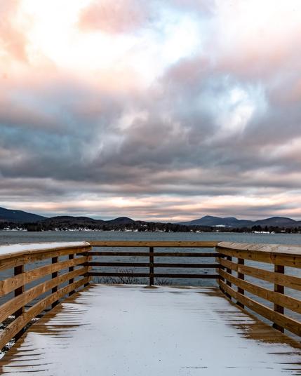 Looking from a snow covered wood boardwalk at a lake with mountains in the background. In the sky the clouds are dramatic with orange and pink highlights