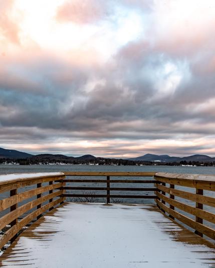 Looking from a snow covered wood boardwalk at a lake with mountains in the background. In the sky the clouds are dramatic with orange and pink highlights