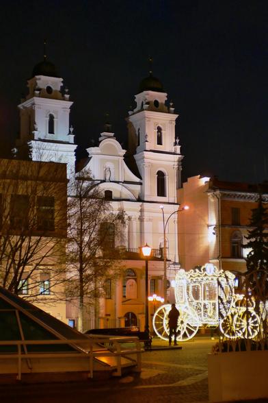The Catholic Cathedral of Saint Virgin Mary, at night in October 2025, viewed from the Town Hall Square in Minsk, Belarus. A glass pyramid on the Town Hall square is visible on the left, and a shining decorative chariot with the coat of arms of Minsk at the top is at the right. A person is standing in front of it.