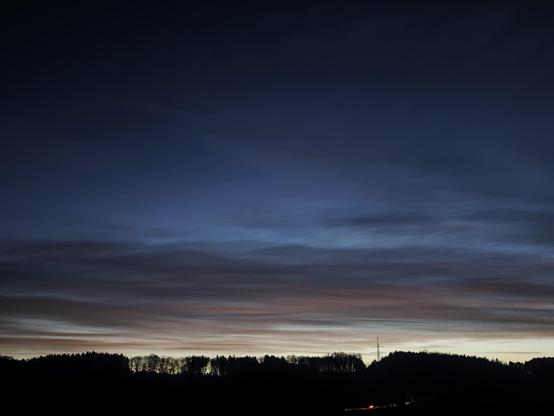 Das Foto zeigt einen dunkelblauen bis schwarzen Himmel mit langgezogenen Wolken, die im Farbverlauf zur bald aufgehenden Sonne von grau über zart-rosa-orange zu hellgold schimmern. Am Horizont ist die Silhouette eines Waldes zu sehen, der sich dunkel vom Himmel abhebt. Im unteren Bereich des Bildes sind schwache Lichter eines Autos erkennbar.