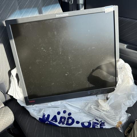 A black LCD monitor labeled Lenovo ThinkVision is perched in a car seat, on top of a plastic bag labeled HARD OFF. The monitor has a price tag that says 550 yen. There are also some RJ11 phone cables