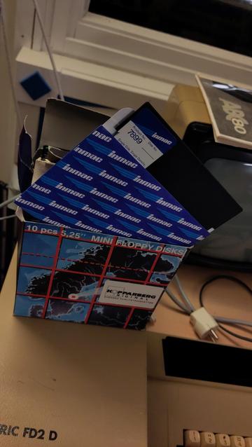 Close-up of a carton box with a 5.25" black rectangle in a paper sleeve standing diagonally on its edges. There's a small label on the top left corner of the black rectangle which says 'inmac', 'product no. 7899', and 'Single Side, Single Density, Soft Sector'.