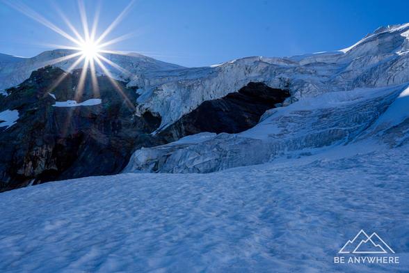 Sunstar shining above a glacier with deep blue ice and rugged textures, the light reflecting off the snow and creating a bright, crisp alpine atmosphere.