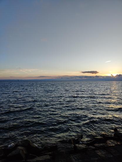 Blick von Romanshorn Hafen Richtung Osten über den Bodensee, über den Vorarlberger Alpen geht rechts die Sonne auf, rosa und orange ein schmales Wolkenband darunter vor den Bergen, das andere Ufer nur dunkel zu sehen, leichter Wellengang, blaues und dunkles Wasser, links die Leuchtturmdalbe, im Vordergrund Steinblöcke der Uferbefestigung