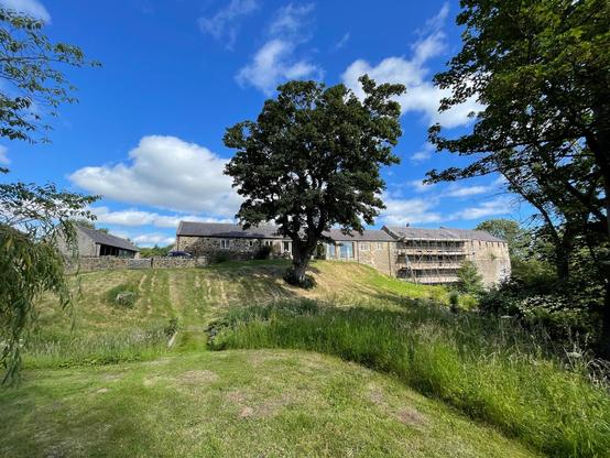 A stone farmhouse complex sits on a gentle grassy slope under a bright blue sky with scattered clouds. The main building features multiple pitched roofs, with part of the structure covered in scaffolding indicating ongoing work. A large tree stands prominently in the center of the slope, casting some shade over the area. Several smaller stone outbuildings lie to the left, partially enclosed by a low stone wall. The surrounding area is a mix of short-cut grass and taller wild vegetation, with trees framing the right side of the scene. A parked car is visible near the main building, and the overall setting is quiet and rural.