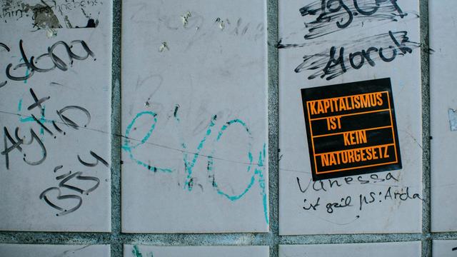 Mehrere handgeschriebene Graffiti und ein schwarzer Aufkleber mit orangefarbener Schrift 'KAPITALISMUS IST KEIN NATURGESETZ' auf einer grau gefliesten Wand. (Foto von Markus Spiske auf Unsplash)
