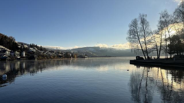 Lake surrounded by hills and a few leafless trees illuminated by the newly risen sun with a few low-lying clouds in an otherwise blue sky.
