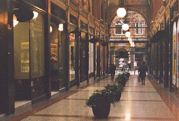 The image shows an elegant Victorian shopping arcade with warm, dim lighting and richly decorated architecture. A row of glowing spherical lamps hangs from the high ceiling, drawing the eye toward an ornate archway at the far end. Polished shopfronts line both sides, their glass reflecting the lights. Large potted plants sit in a neat row along the center of the tiled floor. A few people walk toward and away from the exit, where daylight pours in and silhouettes them slightly. The overall scene feels nostalgic, intimate, and atmospheric, with the texture of film grain adding a softly vintage mood.
