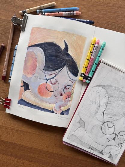 An illustrative drawing of a character with glasses and rosy cheeks is displayed on a white page, accompanied by a sketch in pencil on a nearby page. Various colored pens and pencils are scattered around on a wooden surface.
