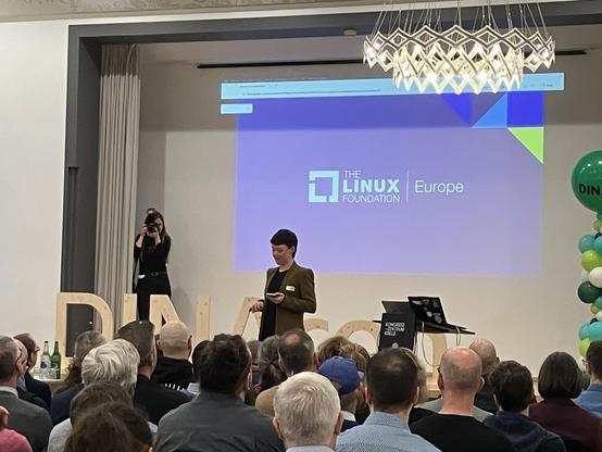 Speaker in front of a slide „The Linux Foundation Europe“ at a conference with photographer in the background and an audience