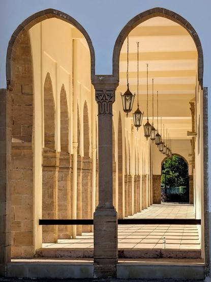 A photograph captured through a symmetrical double archway of a long, sunlit arcade or portico. The arches frame a central, fluted column. The arcade features numerous stone columns with repetitive arches receding into the distance, creating strong perspective lines. A row of ornate, traditional lanterns hangs from the ceiling. The floor is covered in long, distinct shadows cast by the columns and arches. Green foliage is visible at the very end of the corridor.
