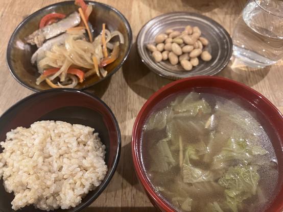 A meal featuring a bowl of brown rice, a bowl of soup with leafy greens, a side dish of cooked vegetables and fish, and a small plate of soy beans. Water is visible in a glass beside the food.