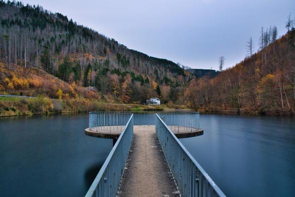 Das Bild zeigt eine ruhige, landschaftliche Szene, wahrscheinlich an einem See oder Stausee in einer bergigen oder hügeligen Region.
Vordergrund: Im Zentrum des Vordergrunds befindet sich ein schmaler Steg mit einem einfachen Metallgeländer, das von unten in die Mitte des Bildes führt. Der Steg endet in zwei halbkreisförmigen Plattformen mit dem gleichen Geländer, die über dem dunklen Wasser des Sees liegen. Die Perspektive führt den Blick direkt über den Steg in die Ferne.
Mittelgrund: Der See oder Stausee dominiert den Mittelgrund. Das Wasser ist dunkelblau bis fast schwarz und erscheint sehr ruhig, was auf eine Langzeitbelichtung hindeuten könnte, die die Oberfläche glättet. Am gegenüberliegenden Ufer, genau in der Mitte des Bildes, steht ein kleines, weißes Gebäude, das von Bäumen umgeben ist.
Hintergrund & Landschaft: Die Hänge beider Seiten des Tals steigen steil an. Die Vegetation zeigt eine Mischung aus Nadel- und Laubbäumen. Es ist wahrscheinlich Herbst, da viele Laubbäume am linken Hang gelbe und orangefarbene Farbtöne aufweisen. Auf dem rechten Hang und an den höheren Stellen sind einige Bäume kahl oder abgestorben, was der Szene eine leicht kühle oder raue Atmosphäre verleiht. Links ist auch ein Abschnitt einer Straße oder eines Weges sichtbar.
Stimmung: Die Beleuchtung ist gedämpft und diffus, möglicherweise ein später Nachmittag oder ein bewölkter Tag. Die gesamte Szene strahlt eine Stimmung der Stille und Isolation aus, wobei der Steg als Verbindungspunkt zwisc