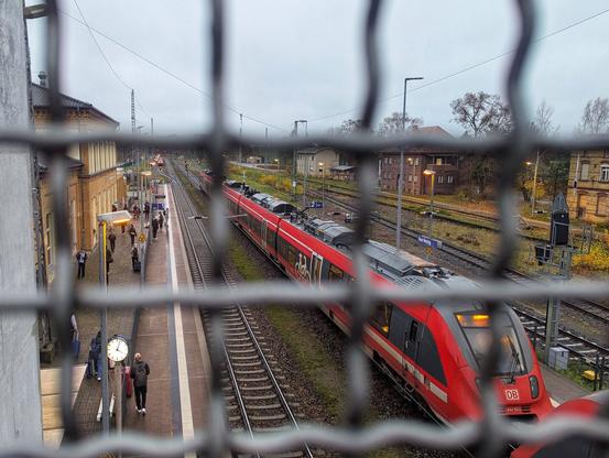 A view through a metal fence shows a train platform with a red Deutsche Bahn train stationed on the tracks. Passengers are waiting on the platform, some walking around. The platform is lined with overhead wires, signs, and a clock. In the background, there are buildings and trees under an overcast sky, giving the scene a slightly muted, autumnal atmosphere.