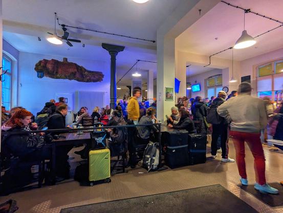 Inside the station, a busy waiting area filled with travellers. People are seated at tables, standing in queues, or walking around with luggage, including suitcases and backpacks. The room is brightly lit with modern lighting, and there is a large, artistic wall decoration of a castle or landmark. The atmosphere is lively, with a mix of individuals and groups.