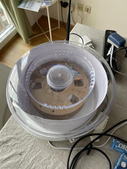 a round food dehydrator with a transparent lid. Under the lid a cardboard spool of 3D printer filament can be seen. the dryer is sitting on a desk that looks slightly messy with various cables and gadgets.