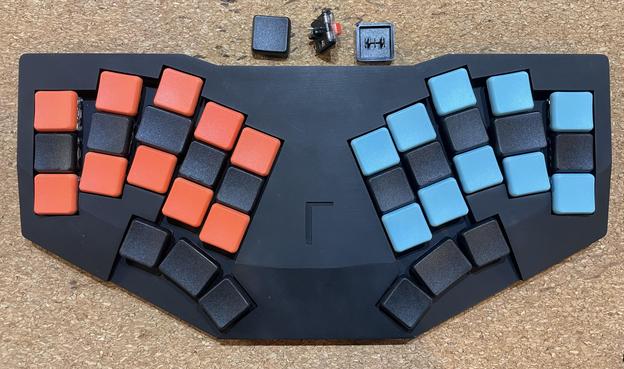 Small 36-key mechanical keyboard in a black resin 3D printed case with Chosfox CFX square keycaps in black (home row and thumb keys), orange (top and bottom on left), and robin blue (top and bottom on right). The middle thumbs have a 1.25u keycap, as do the S&L keys (could do this at D&K instead), which fit because these are slightly smaller than the Kailh Choc keycaps this case was designed for. The index and pinky home keys have a homing bumb.