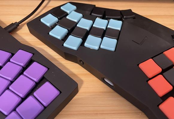 Two small black keyboards, on the left with a USB cable the wired only Gamma Omega TC36K (with purple MKB keycaps), and on the right showing a reset button and USB port on the top edge, the wireless Gamma Omega Hesse (with blue, black, and orange CFX keycaps).