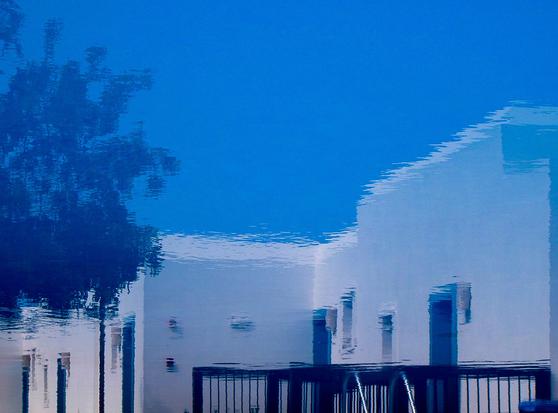An abstract reflection of a building and trees on a water surface, creating a distorted image against a blue sky backdrop.