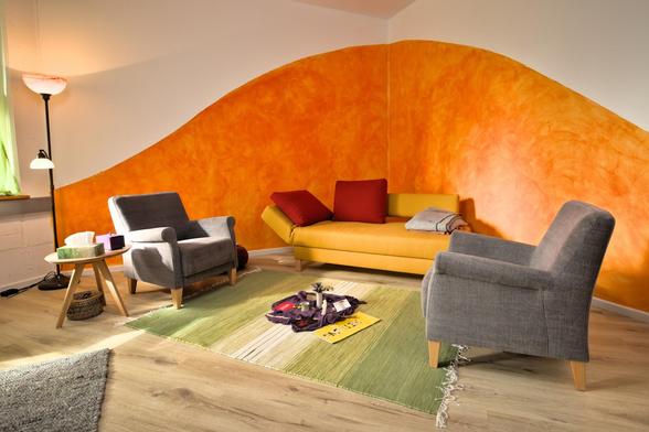 Mein Praxisraum in Eckernförde mit gelbem Sofa mit roten Kissen, zwei grauen Sesseln auf einem hellgrünen Teppich und einer orange gestrichenen Wand im Hintergrund. Klein zu sehen ist die gestaltete Mitte auf dem Teppich mit rituellen Gegenständen.