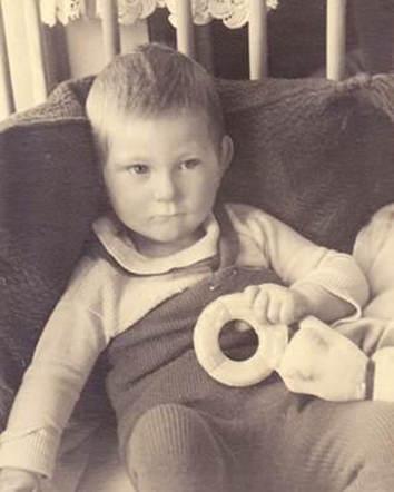 A little boy with short blonde hair lies on a bed leaning against a pillow (the rungs are visible in the background). In his left hand he holds a toy - a lifebelt.