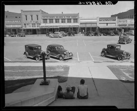 The black and white photograph depicts a bustling small-town American downtown with several antique cars parked in front of various stores. There are at least seven vintage automobiles from the 1930s era, identifiable by their distinct features such as rounded fenders and large bumpers.

In the foreground, three men sit on steps looking out onto the street; two wear hats while one has a white shirt with dark pants. The architecture suggests early to mid-20th-century design, characterized by flat facades and decorative signage above storefronts advertising goods like groceries, jewelry, body glass, hamburgers, and more.

The sky is clear indicating it might be a sunny day as evidenced by the sharp shadows cast on the pavement. There's no visible traffic signal or pedestrian crossing in sight which was common during that era for safety reasons. The overall mood of the photograph captures everyday life from what appears to be an ordinary town square, reflecting societal norms and urban planning typical of small American towns at the time.

This image is a visual representation captured by Dorothea Lange as part of her extensive work documenting various aspects of American life during the Great Depression.