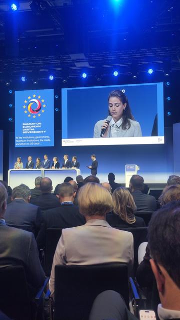 Ein öffentliches Treffen findet in einem Konferenzraum statt. Auf der Bühne steht eine Person mit braunen Haaren und einem blauen Nadelstreifenanzug und hält ein Mikrofon fest. Unter der Person auf der Bühne befinden sich mehrere Reihen von Personen, die alle nach vorne schauen. Im Hintergrund befindet sich ein Bildschirm mit dem Text „SUMMIT ON DIGITAL SOVEREIGNTY“ darüber „Its key institutions, governments, healthcare institutions, the media and others“. Auf dem Bildschirm befindet sich außerdem ein Logo mit einem Sternmuster.

Danke, Altbot