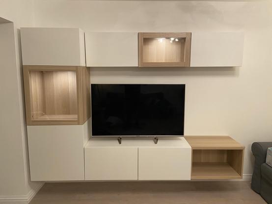 Empty Ikea Bestå cabinets mounted on the wall. It is in a C-shape of 4 units wide mostly in white with solid white doors. The left hand column has two taller units and a shorter one on top with the middle unit being glass fronted and oak effect framed. To the right of the left column are three shorter units at the top and bottom with the top row being shallower. The centre of the three shallower top units is also glass fronted and oak effect framed. The right most of the bottom units is also oak effect but with no door.