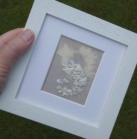 Stitched artwork on hand dyed fabric in pale lilac with a lighter, tye dyed starburst shape. The stitching comprises an abstract shape, somewhat resembling a prawn, made up of triangles in very shiny, white and grey silk thread. Framed in a 5 x 5 inch white frame and held in the artist's hand for scale