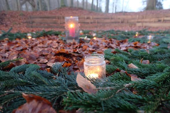 eine Kerze im Glas im Vordergrund auf grünen Tannennadeln, im Hintergrund eine Kerze in einem größeren Glas; beide Kerzen stehen im Wald.