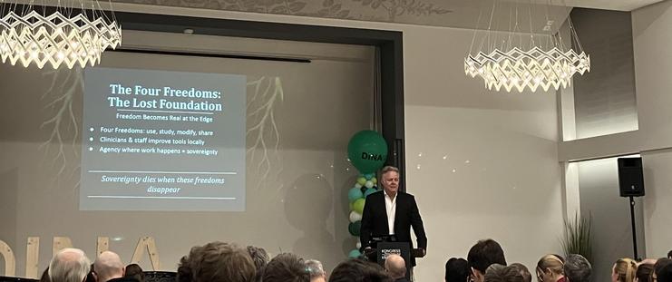 A speaker on stage in front of green balloons and a large audience. Transcript of slide:

The Four Freedoms:
The Lost Foundation

Freedom Becomes Real at the Edge
• Four Freedoms: use, study, modify, share
• Clinicians & staff improve tools locally
• Agency where work happens = sovereignty

Sovereignty dies when these freedoms
disappear