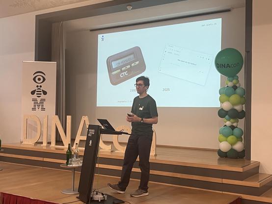 Photo by Kate Schütz (CC BY 4.0)

A speaker on the stage with a slide showing an old fashioned pager device on one side and an AI prompt window on the other.