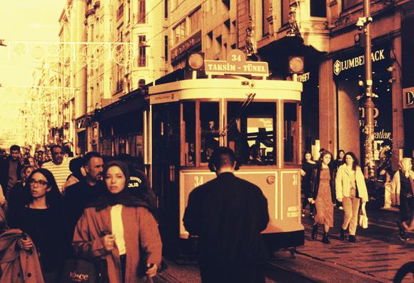 A busy city street captured on redscale film, featuring a classic red tram moving through crowds of pedestrians between tall storefront-lined buildings.