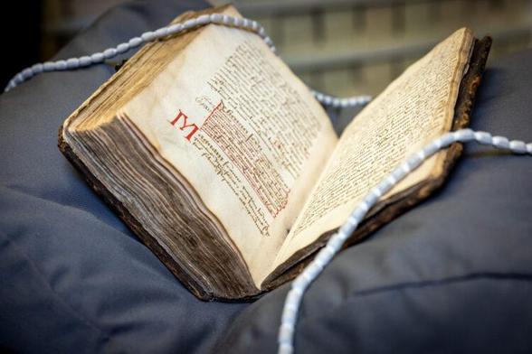 A sixteenth-century handwritten book, lying open on a dark grey-blue cushion. Cloth-covered weighted cords hold the book open to display the text, including a section underlined in red and a paragraph starting with a large, red capital M.