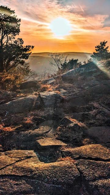 Coming out onto a cliff of coarse brown rock with numerous cracks and fissures. There are patches of golden brown dormant grass growing in many of the cracks. A forest of cedars and leafless hardwoods grows on either side of the cliff. The view is just appearing, some rolling hills and buildings a few hundred feet below. The late afternoon sky is a pale blue gray. Most of the visible sky is covered with a cluster of hazy thin clouds. The sun, setting behind the haze, is a large roundish blob of golden fire, painting the surrounding haze orange and yellow.