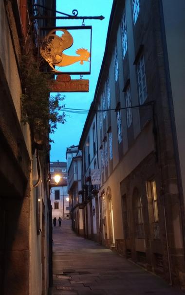 Narrow cobblestone alley at dusk with warm streetlights. A decorative sign with a golden lion hangs on the left. Two people walk in the distance.
#laureanoaraujo #santiagodecompostela #galiza
Santiago de Compostela, 18 de novembro de 2025