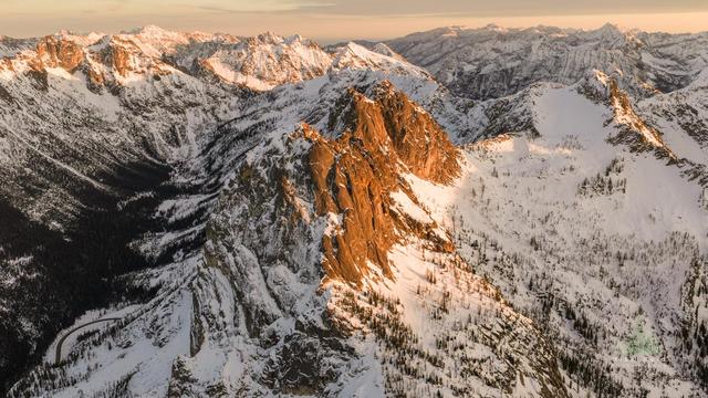 This aerial landscape captures Liberty Bell Peak glowing in warm sunset light above Washington Pass in the North Cascades. Evening alpenglow spreads across the rugged granite walls and snowy ridgelines of the Liberty Bell group.