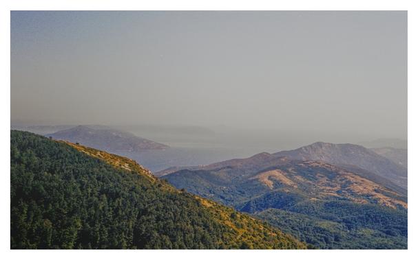 Color photograph of the Adriatic Sea and Croatian islands, taken from Vojak, the highest peak in Istria and the Učka Mountains. In the foreground, densely wooded hills can be seen stretching toward the horizon in lower hills and eventually merging with the sea. The sky swallows the hills on the horizon because it was so hazy.