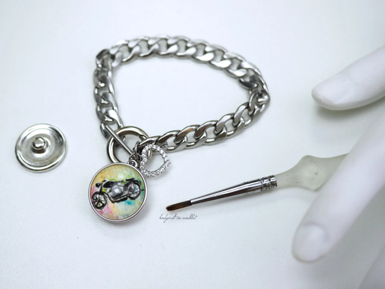 handgemalter Druckknopf-Motivbutton mit stilisierter schwarzer filigran gemalter Motorradsilhouette auf mehrfarbigem Grund an 30 Gramm schwerem, massive Edelstahl-Gliederarmband mit einem Charm mit Druckknopfhalterung und einem weiteren Charm mit kleinem Herz mit Glassteinen