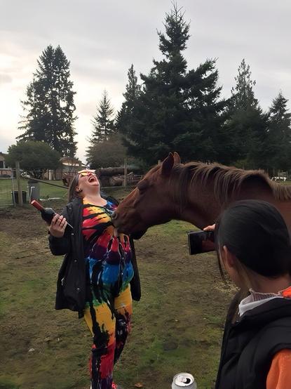 A brunette woman in a tie-dye onesie holding a wine bottle is being licked by a brown horse, with me, in a fox onesie, taking a photo with my phone. The brunette is laughing with her head tilted back, wearing red-framed sunglasses. The woman has a black jacket over rainbow tie-dyed onesie. The horse is extending its head towards her chest & licking. The background includes cloudy skies, trees, and a farm field with a fence.
