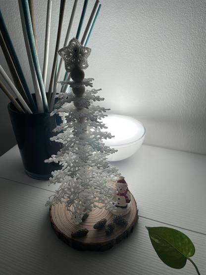 Albero di Natale fatto ad uncinetto con filo bianco su base di legno, vicino ci sono un pupazzetto di neve e delle pigne. 
A fianco c’è un contenitore con bastoncini profumati e una luce colorata. In basso spunta anche una foglia. 
Tutto su tavolino bianco.