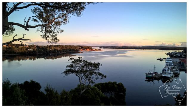 A wide, horizontal view of Macquarie Harbour in Strahan, Tasmania, at sunset. The calm water reflects the soft gradient of the sky, which transitions from pale blue at the top to a warm orange pink near the horizon.  In the foreground, dark, lush foliage and a single small tree on an outcrop frame the scene, with the silhouette of a large tree branch extending over the upper left.  To the right, several fishing boats are moored near a dock and a cluster of low buildings.