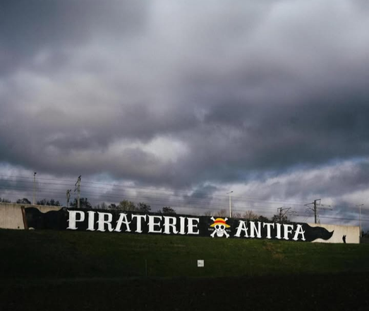 A large graffiti saying “piraterie antifa” on a concrete wall. It has a black background and that pirate flag from one piece in between the two words