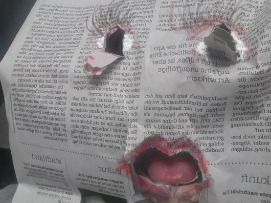 Zeitungsblatt vor Gesicht, auge- und mundlöcher reingerissen. In dem Loch für den Mund sieht man echte Zunge. Lippen und augenbeteich sind auf die Zeitung gemalt.