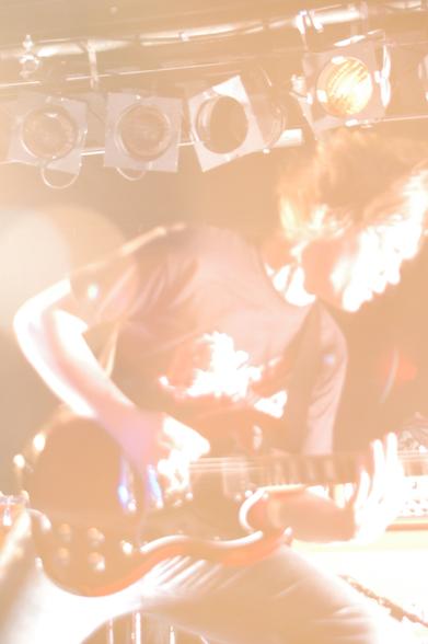 A massively over-exposed and pretty-much blown-out photo of a man with long, light-coloured hair, wearing a black t-shirt with a white logo, and playing a red electric guitar.
Bright stage lights, some mounted on rails at the top of the walls in the background, have flooded the frame with an explosion of orange light, washing out a lot of the detail, yet leaving enough to form a dynamic image.