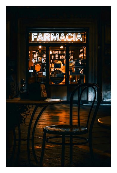 A dimly lit, cozy looking pharmacy at night. The neon sign "FARMACIA" glows brightly above the glass door. Inside, warm orange lights illuminate shelves of bottles and a counter. In the foreground, a black, wickered chair with a curved backrest sits at an empty, round wooden table. The glow of the lights are reflected on the damp street cobbles.
