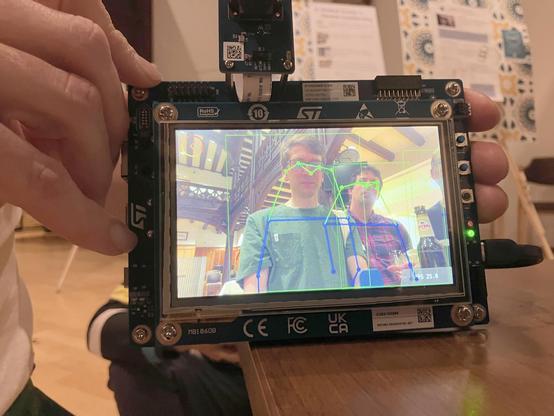 Photo of a hands holding a display with a webcam feed with pose tracking of two people. Part of the WildCamera project.