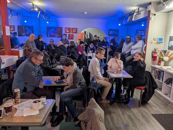 People packed around tables and standing in a Small club.  There is almost no free space to move around.