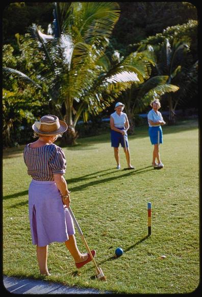 In this photograph from Toni Frissell's collection, three individuals are engaged in a game of croquet on the grass. The prominent figure is an older woman with her back to the camera, wearing a straw hat and holding a mallet aimed at a red-painted hoop near the center-right side of the image. She dons a striped blouse paired with a purple skirt and pink sandals.

Behind this player are two other individuals observing the game; one appears readying for their turn while dressed in blue shorts and white shirt, and the other is standing slightly further away wearing similar attire. The scene unfolds on well-kept grass amidst an environment richly adorned by palm trees that tower above them under a bright sky.

Additional context reveals this image was captured at Mill Reef Club located in Antigua during January of 1959 as indicated by the reference information provided, which also highlights Toni Frissell's professional involvement with Sports Illustrated between 1959 and 1964.