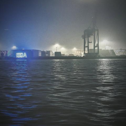 In einer nebligen Hafenlandschaft erhebt sich ein gewaltiger Containerkran, der im diffusen Licht der umgebenden Laternen mystisch wirkt. Der Vordergrund wird von sanften Wellen des Wassers dominiert, die das Licht reflektieren und eine ruhige, fast melancholische Stimmung erzeugen. Auf der linken Seite leuchtet ein Container in einem intensiven Blau, das den Schriftzug "HHLA" trägt und einen modernen Akzent setzt. Die Szenerie ist in gedämpften Grautönen gehalten, die durch das Nebellicht verstärkt werden, während die Lichter im Hintergrund eine geheimnisvolle Atmosphäre schaffen. Die Komposition vermittelt eine Mischung aus industrieller Stärke und stiller Gelassenheit, die den Betrachter in eine Welt der Hafenarbeit und des nächtlichen Transports entführt. [KI-generiert]